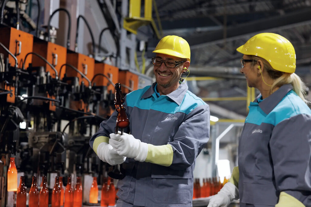 Eine Mitarbeiterin und ein Mitarbeiter in der Glasproduktion bei Verallia, tragen Schutzhelme und Arbeitskleidung, der Mann lächelt und hält eine frisch geformte Glasflasche.